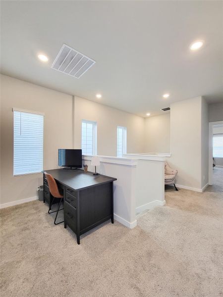 Bright, carpeted room with recessed lighting, featuring a small office area with a desk and chair. There are three windows allowing natural light to enter, and an additional seating area is visible in the corner.