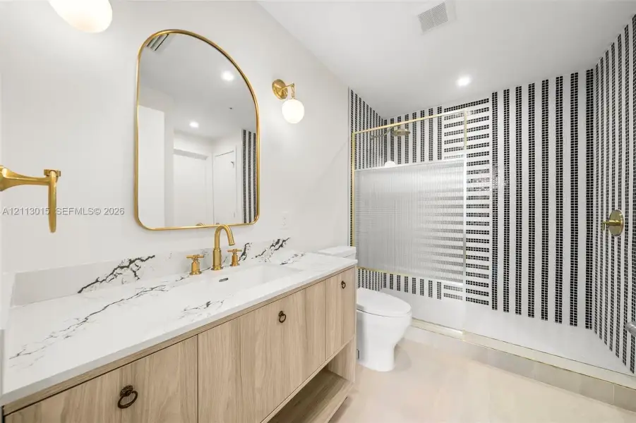 Vanity with wood-finish cabinetry, stone countertop with veining, arched wall mirror, gold-toned fixtures, and a shower enclosure featuring vertical striped mosaic tiling