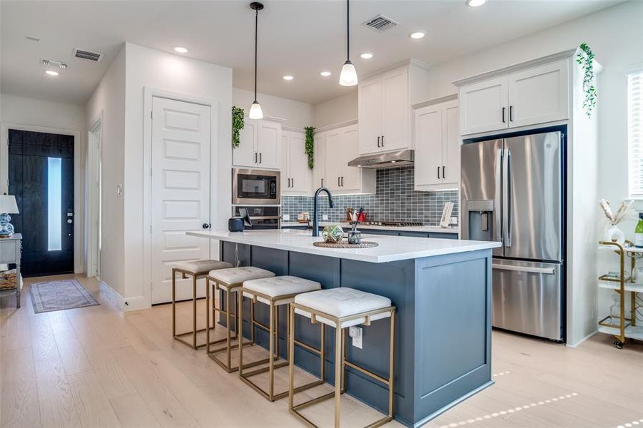 Two tone kitchen with stainless steel fridge with ice dispenser, an island with sink, two tone cabinetry, light wood finished floors, and decorative light fixtures