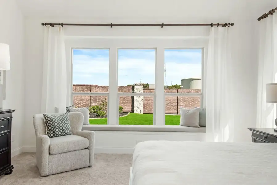 Bedroom with window seat, gray armchair, white curtains, and backyard view through triple windows