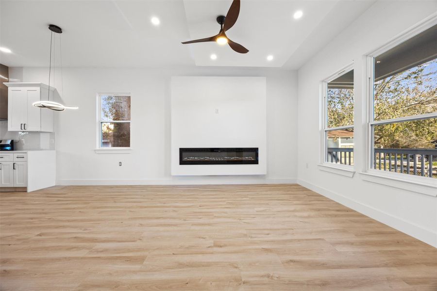 This spacious living area features light wood-like flooring, large windows for natural light, and a modern recessed fireplace. The open layout flows into the kitchen with sleek cabinetry, enhanced by contemporary ceiling fans and lighting. Perfect for a bright, inviting home environment.