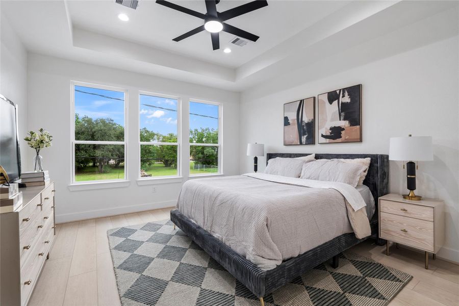 Primary bedroom with tray ceiling, modern fan, and a wall of windows overlooking greenery. Primary bedroom with tray ceiling, modern fan, and a wall of windows overlooking greenery.