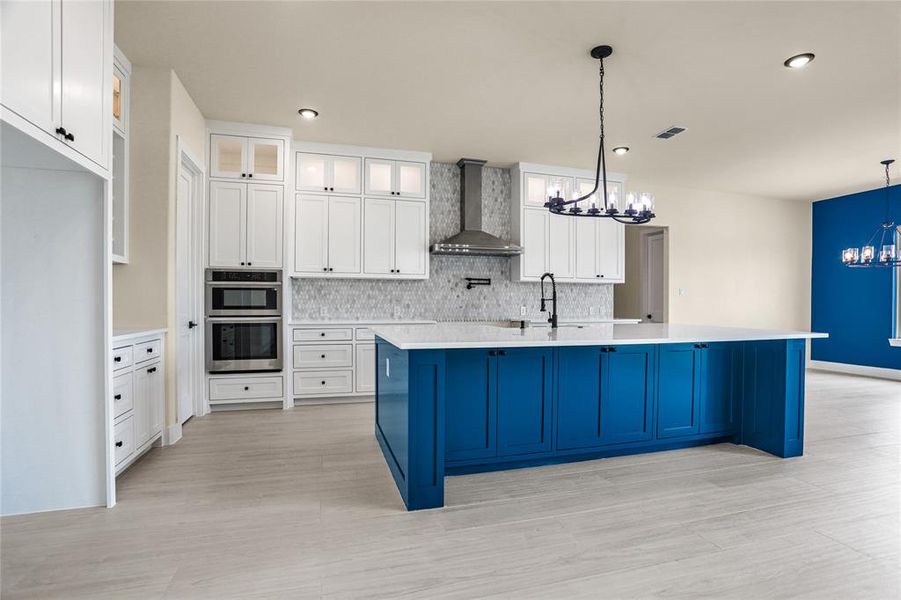 Kitchen with a chandelier, wall chimney exhaust hood, stainless steel double oven, light wood-style flooring, and tasteful backsplash Kitchen with a chandelier, wall chimney exhaust hood, stainless steel double oven, light wood-style flooring, and tasteful backsplash