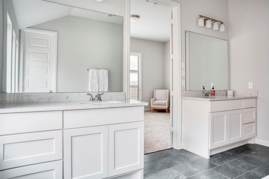 This photo showcases a modern bathroom with dual sinks, white cabinetry, and a large mirror. It features sleek gray tile flooring and an adjoining carpeted room with natural light, creating a bright and inviting space. This photo showcases a modern bathroom with dual sinks, white cabinetry, and a large mirror. It features sleek gray tile flooring and an adjoining carpeted room with natural light, creating a bright and inviting space.