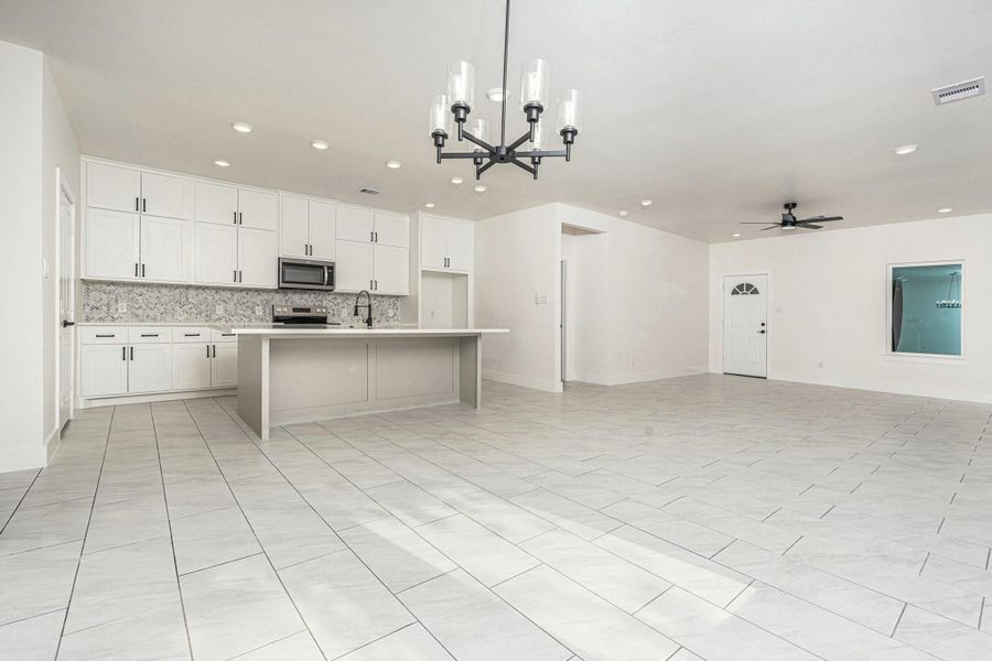 This spacious open-concept area features a modern kitchen with white cabinetry, stainless steel appliances, and a large island. The room has tiled flooring, neutral walls, recessed lighting, and a contemporary chandelier, ideal for entertaining and family gatherings.