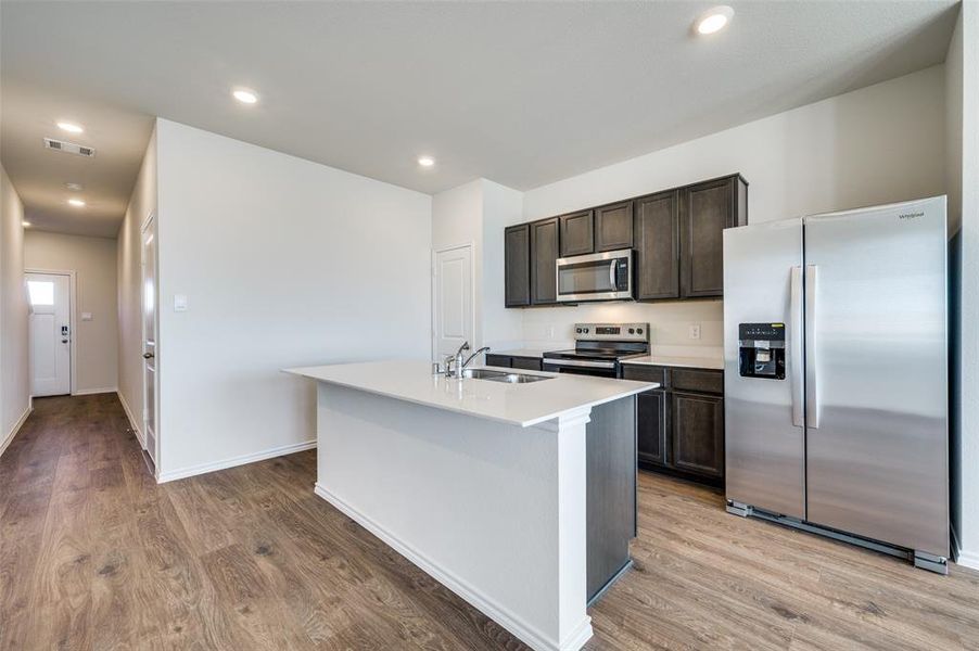 Kitchen with appliances with stainless steel finishes, dark brown cabinetry, a kitchen island with sink, sink, and light hardwood / wood-style flooring