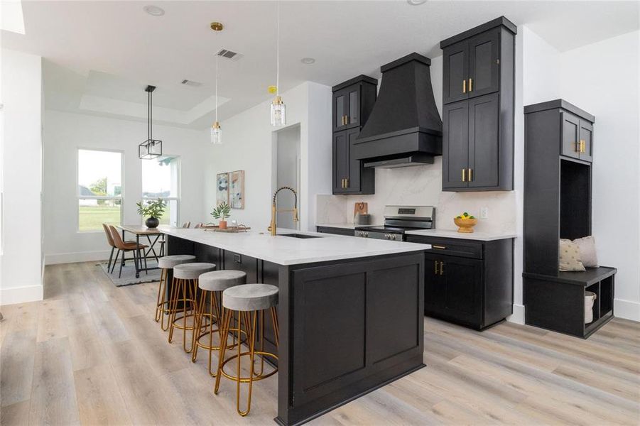 Kitchen with sink, a raised ceiling, an island with sink, stainless steel stove, and custom range hood