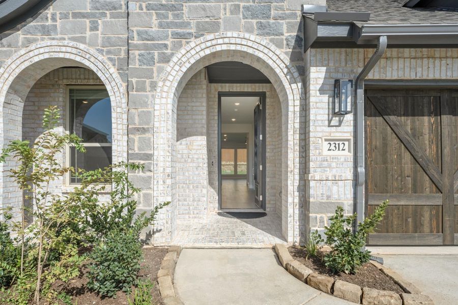 Stone home exterior with arched entryway, gray front door, brick walkway, and wood garage door beside landscaped shrubs