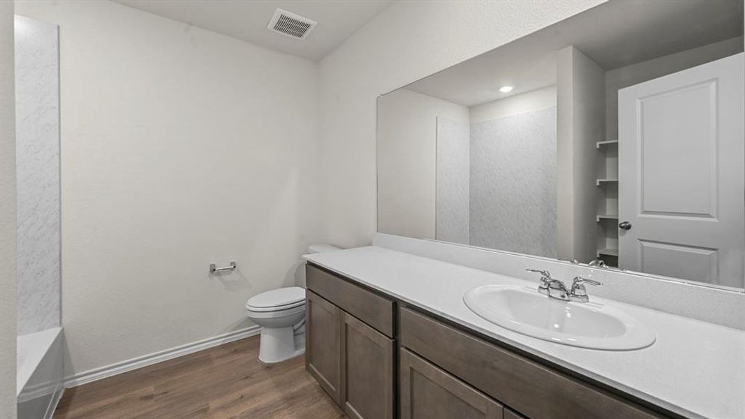 Bathroom featuring a wood-finish vanity with a white countertop and integrated sink, a large wall-mounted mirror, a toilet, a built-in tub with textured white surround, and wood-finish flooring