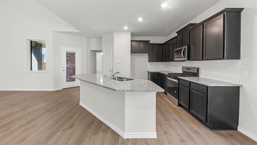 Kitchen featuring appliances with stainless steel finishes, light stone countertops, decorative backsplash, an island with sink, and light wood-style flooring