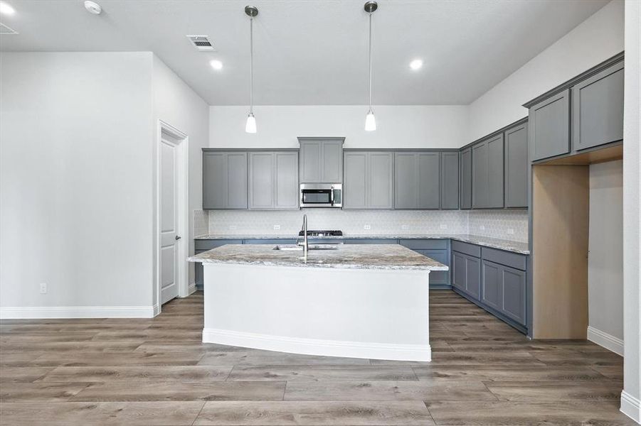 Kitchen with gray cabinets, light stone counters, a kitchen island with sink, stainless steel microwave, and tasteful backsplash Kitchen with gray cabinets, light stone counters, a kitchen island with sink, stainless steel microwave, and tasteful backsplash