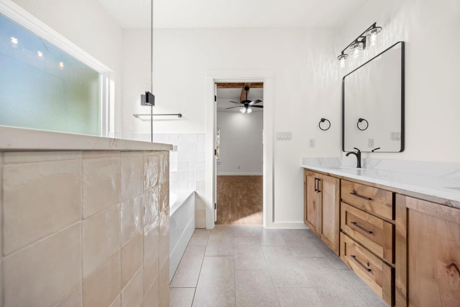 A well-lit bathroom with tile flooring features a bathtub, a vanity with ample storage, and a large mirror.