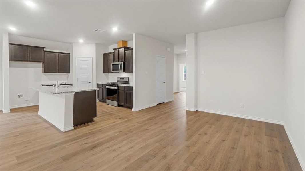 Kitchen featuring appliances with stainless steel finishes, a kitchen island with sink, light stone countertops, dark brown cabinetry, and light wood-style floors Kitchen featuring appliances with stainless steel finishes, a kitchen island with sink, light stone countertops, dark brown cabinetry, and light wood-style floors