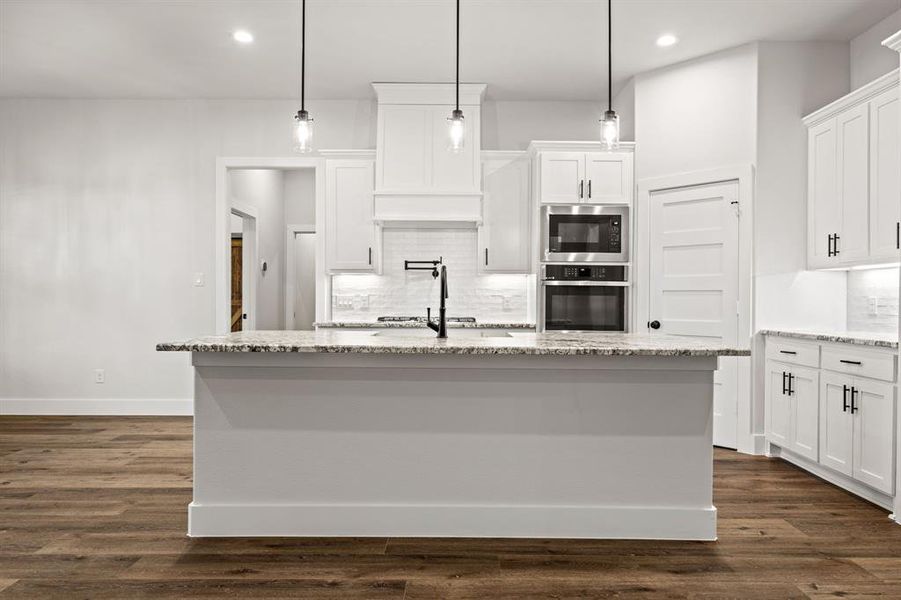 Kitchen with light stone counters, pendant lighting, a center island with sink, built in microwave, and oven