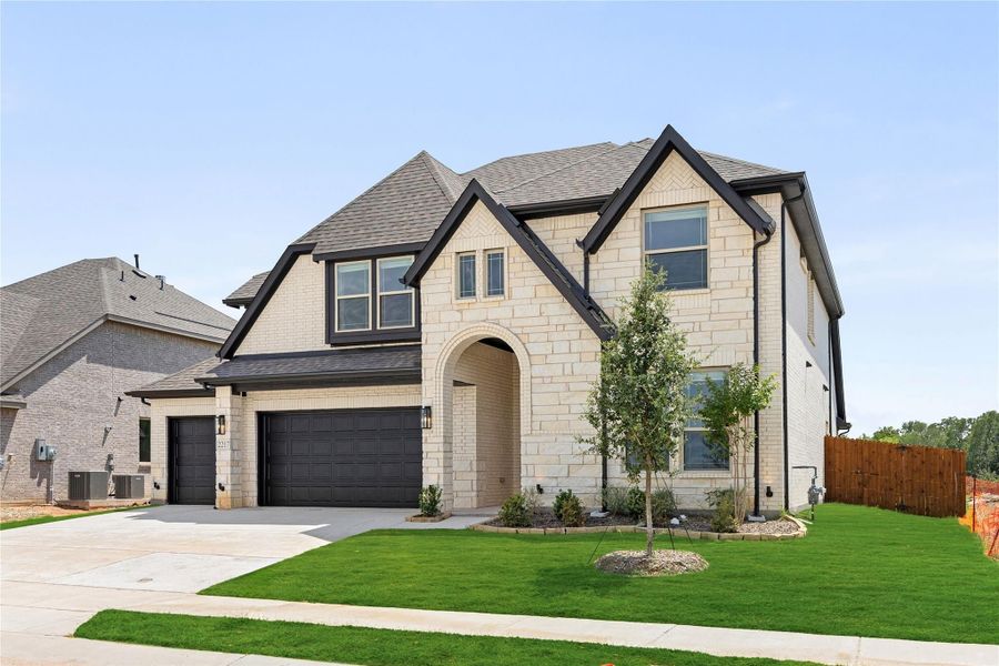 Two-story home with light stone exterior, arched entry, dark two-car garage, and manicured front lawn