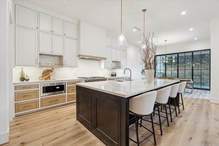 This kitchen is a chef's dream,  showcasing hardwood floors, myriad storage, and dramatic quartzite countertops. Top-of-the-line appliances from Wolf, Sub Zero, and Bosch will elevate your cooking experience. The white backsplash and framing makes the darker tones of white oak drawers and island pop. Soft-close cabinets and drawers with gold pulls further elevate the space.