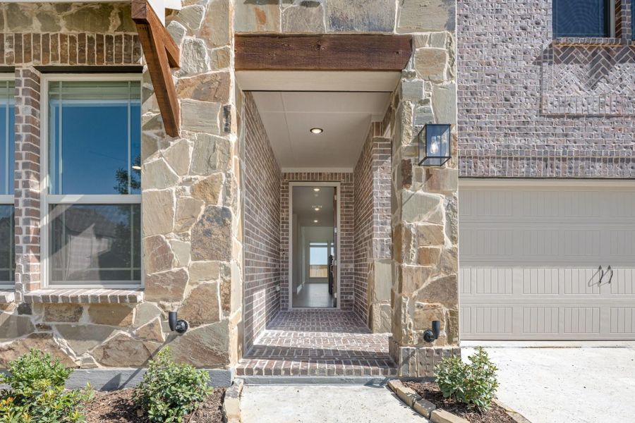 Covered front entry with stone and brick surround, wood beam accent, and wall lantern beside garage Covered front entry with stone and brick surround, wood beam accent, and wall lantern beside garage
