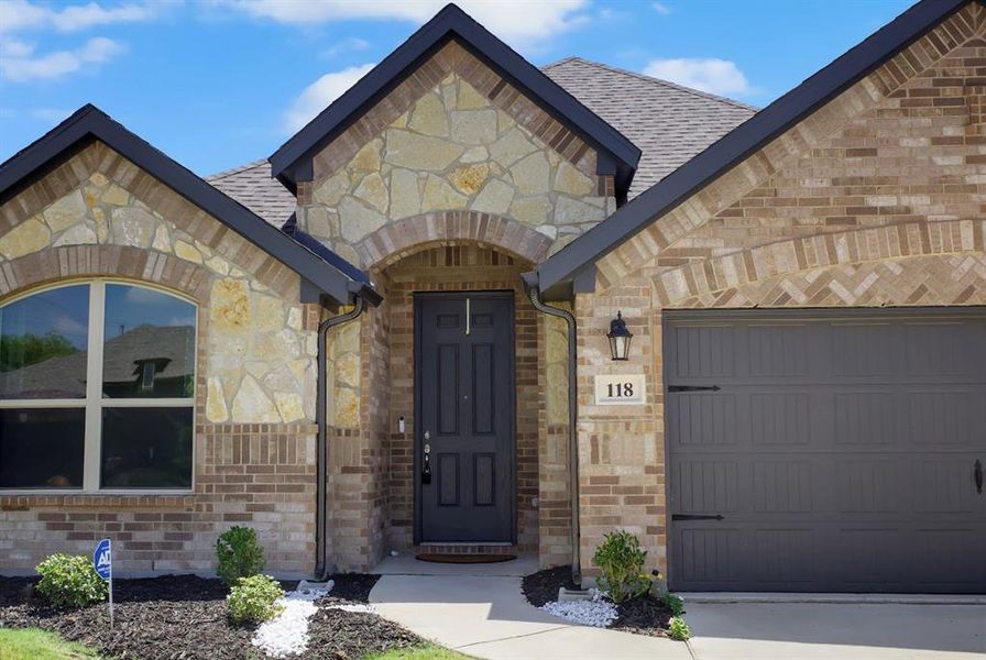 Entrance to property with stone siding, a garage, a shingled roof, and brick siding