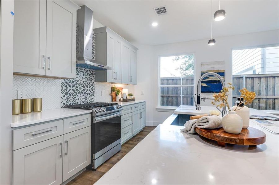 Kitchen featuring wall chimney exhaust hood, stainless steel gas range oven, tasteful backsplash, light stone counters, and decorative light fixtures