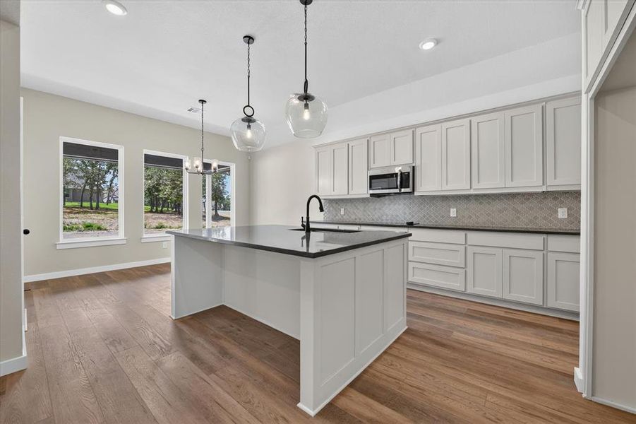 Spacious kitchen featuring a large central island with a sink, extensive white cabinetry, dark countertops, a tiled backsplash, and wood-finish flooring Spacious kitchen featuring a large central island with a sink, extensive white cabinetry, dark countertops, a tiled backsplash, and wood-finish flooring