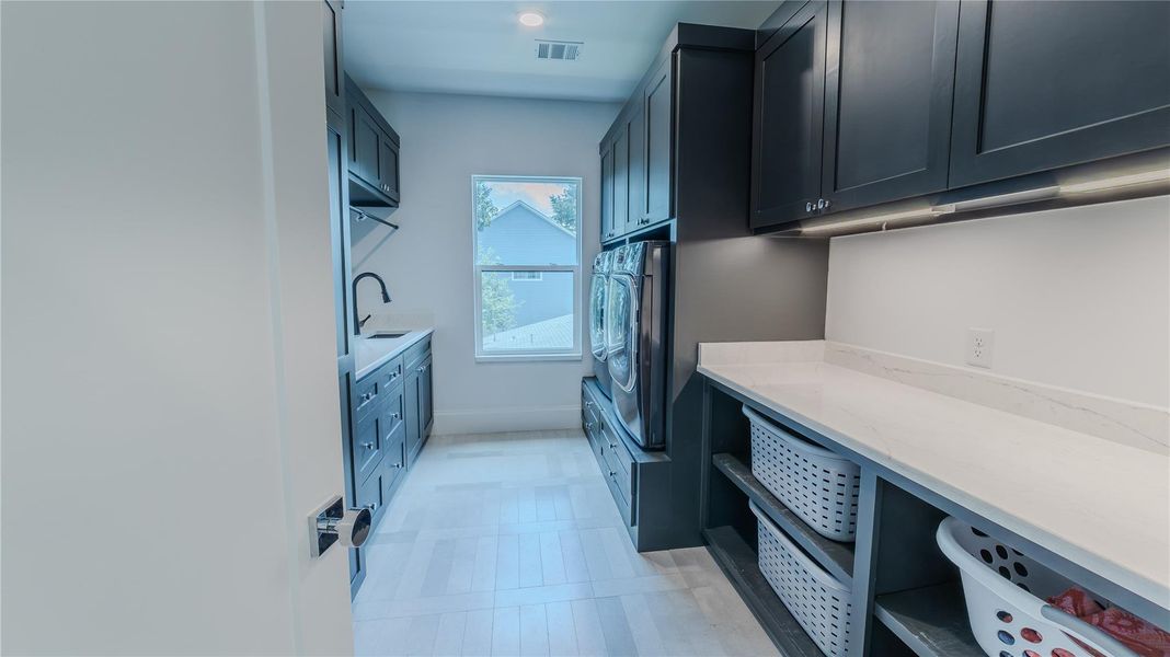 This photo showcases a modern laundry room with sleek dark cabinetry, a washer and dryer, ample countertop space, and storage shelves. The room is well-lit with natural light from a window.