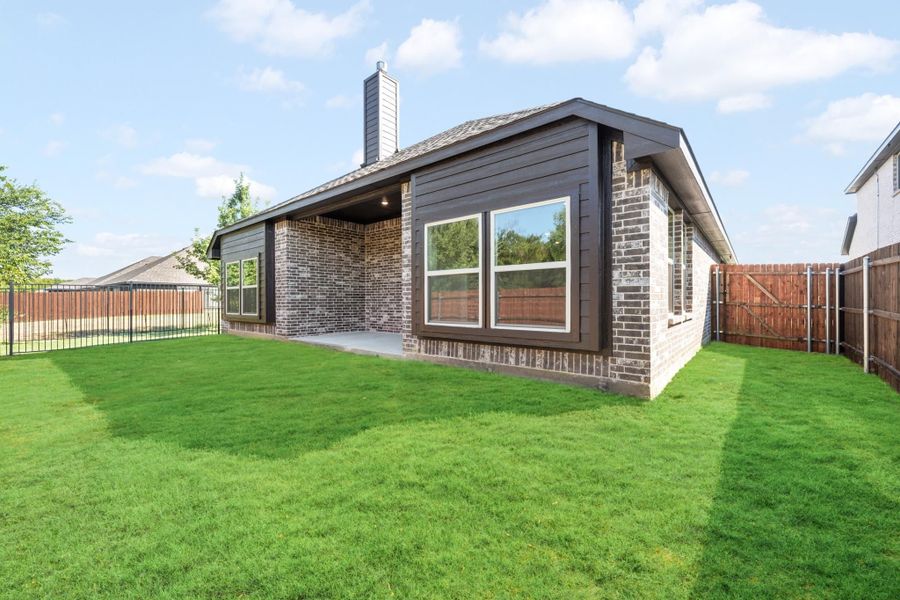 Backyard of single-story brick home with dark siding, covered patio, chimney, and fenced yard with green lawn.