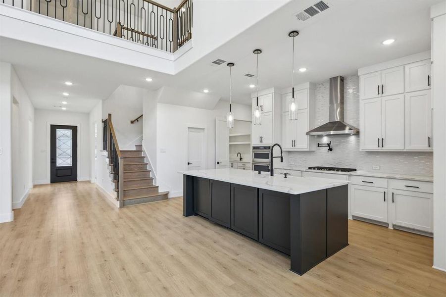 Kitchen featuring white cabinetry, gas cooktop, an island with sink, pendant lighting, and wall chimney range hood