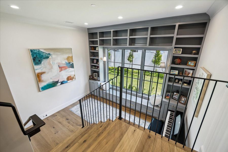 Overlooking the flex space below, the upper landing showcases the home’s open design and impressive scale. Custom built-ins extend to the upper level, adding architectural interest and display space, while the large divided light window fills the area with natural light and views of the side yard and beautiful landscaped grounds.  We love  the view of the privacy wall and landscaping outdoors.