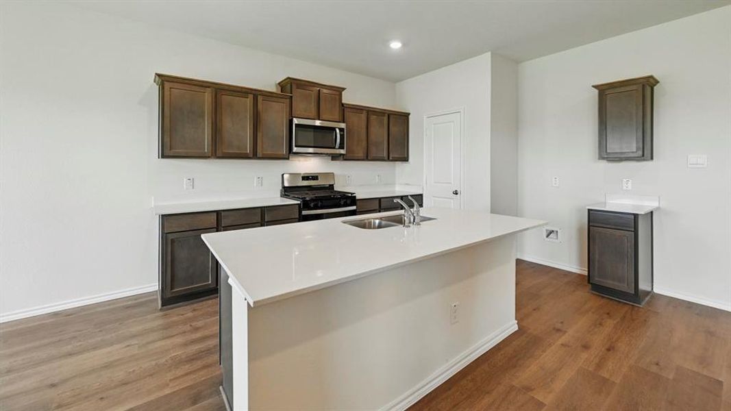 Kitchen with stainless steel appliances, dark brown cabinetry, an island with sink, dark wood-style floors, and light stone counters