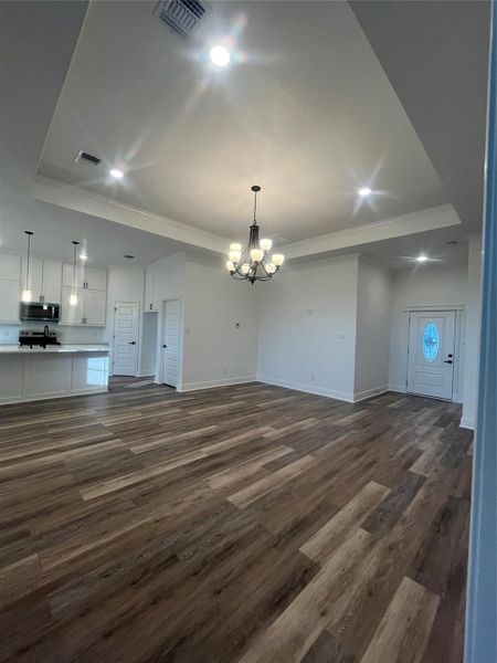 This photo showcases a spacious, open-concept living area with elegant medium gray/wood water proof plank flooring and a modern kitchen. The room features a tray ceiling with recessed lighting and a central chandelier, offering a bright and welcoming atmosphere.