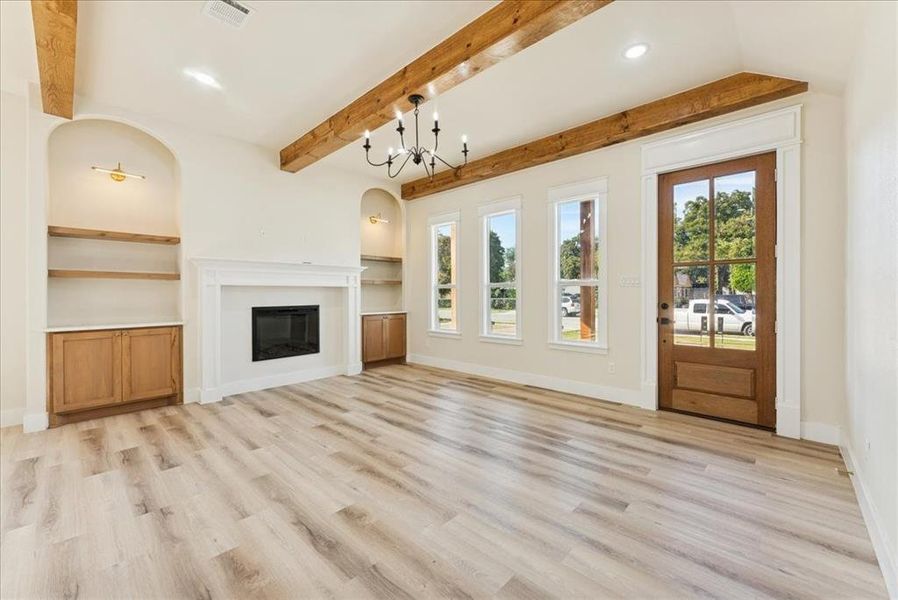 Unfurnished living room featuring a glass covered fireplace, a chandelier, light wood finished floors, beam ceiling, and built in shelves
