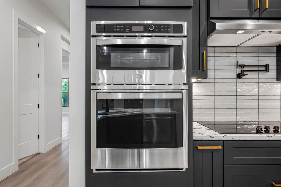 Kitchen with double oven, exhaust hood, tasteful backsplash, black electric cooktop, and light wood-style floors. Kitchen with double oven, exhaust hood, tasteful backsplash, black electric cooktop, and light wood-style floors.