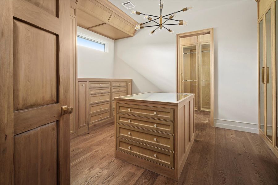 Primary Closet 2 is wrapped in warm custom millwork, with extensive built-in storage, glass-front display cabinetry and a center island with a glass top for showcasing accessories. White oak flooring and a sculptural chandelier complete the tailored dressing space. Primary Closet 2 is wrapped in warm custom millwork, with extensive built-in storage, glass-front display cabinetry and a center island with a glass top for showcasing accessories. White oak flooring and a sculptural chandelier complete the tailored dressing space.
