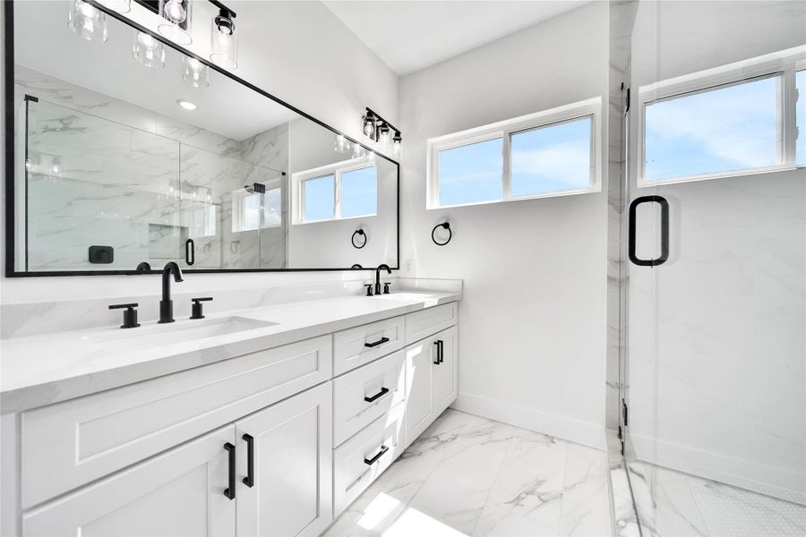 This bathroom features a sleek, modern design with a double vanity, black fixtures, and a large mirror. It has bright, natural lighting from high windows and a spacious glass-enclosed shower, complemented by elegant marble-style flooring and walls.