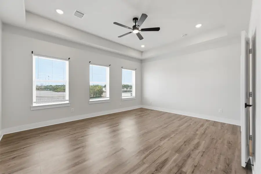 Spacious primary bedroom featuring wide-plank flooring, tray ceiling, modern ceiling fan, and multiple windows providing abundant natural light and expansive views.