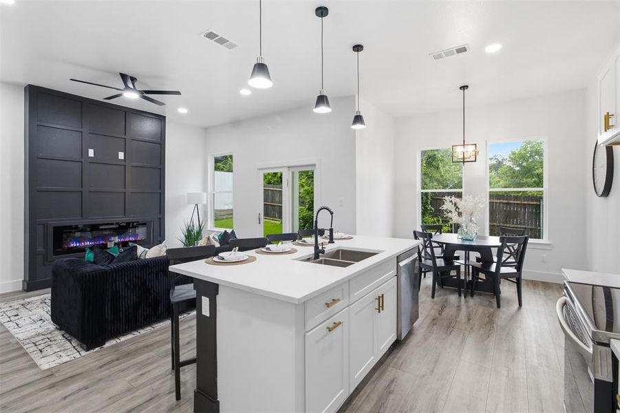 Open-concept interior featuring wood-finish flooring, a kitchen island with a double basin sink, white cabinetry with gold-tone hardware, a paneled fireplace wall, and large windows