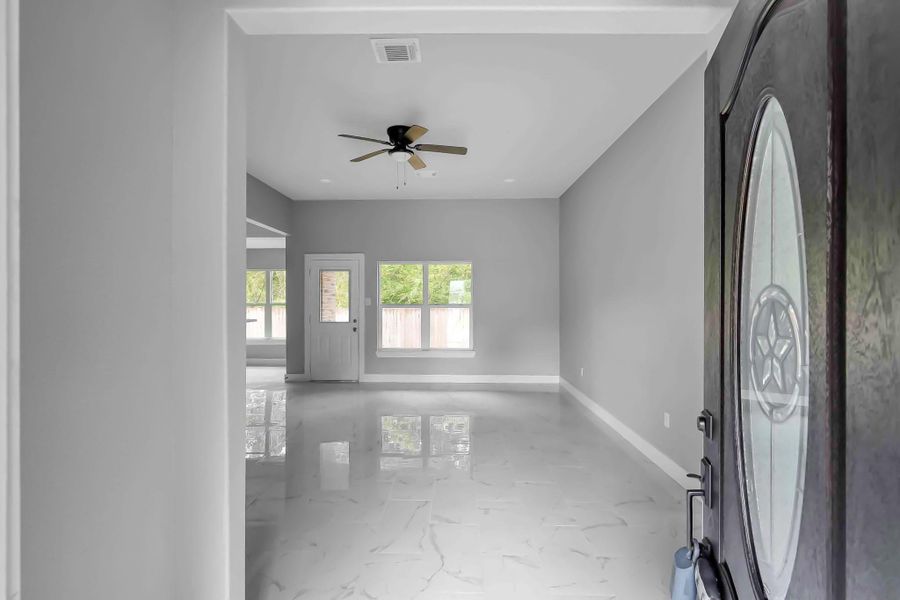 This photo showcases a bright, open living area with glossy tile flooring and neutral gray walls. It features a ceiling fan, large windows for natural light, and a decorative front door.