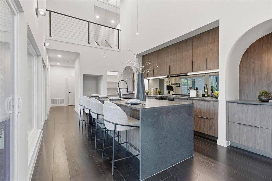 Open-concept kitchen featuring a dark wood-finish flooring, a large island with a stone countertop, contemporary wood-finish cabinetry, built-in shelving, and a second-story interior balcony with metal railings