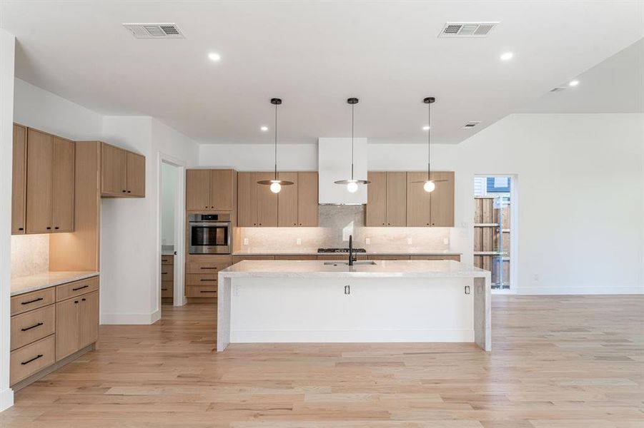 Kitchen with pendant lighting, a center island with sink, oven, light wood-type flooring, and light stone counters