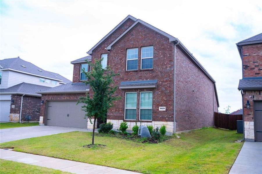 Two-story brick exterior featuring a stone accent foundation, attached two-car garage, and a landscaped front yard with a young tree Two-story brick exterior featuring a stone accent foundation, attached two-car garage, and a landscaped front yard with a young tree
