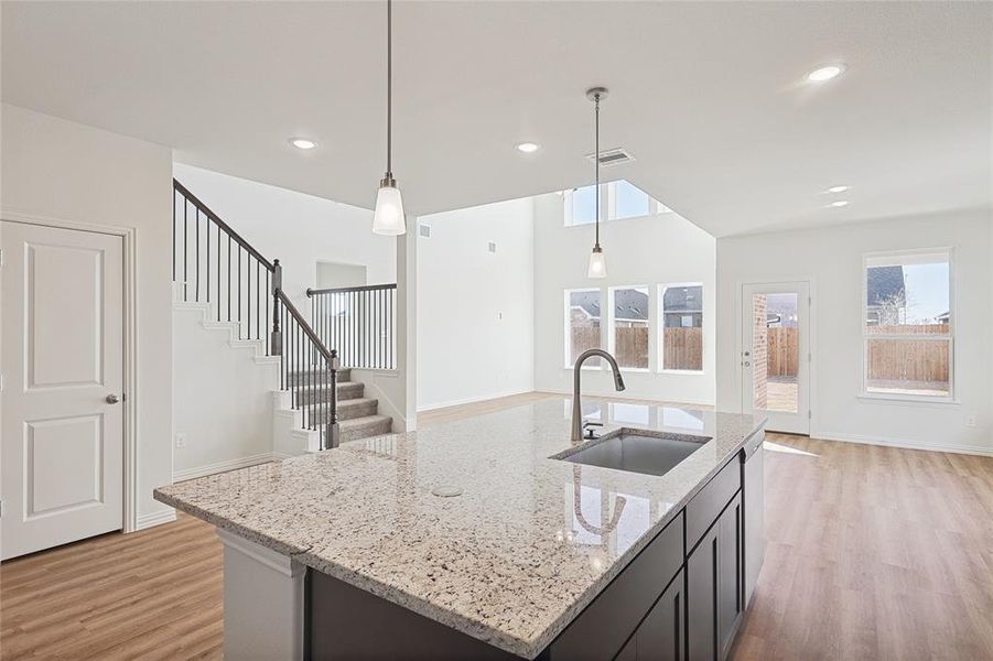 Kitchen with light wood finished floors, light stone countertops, hanging light fixtures, open floor plan, and a kitchen island with sink