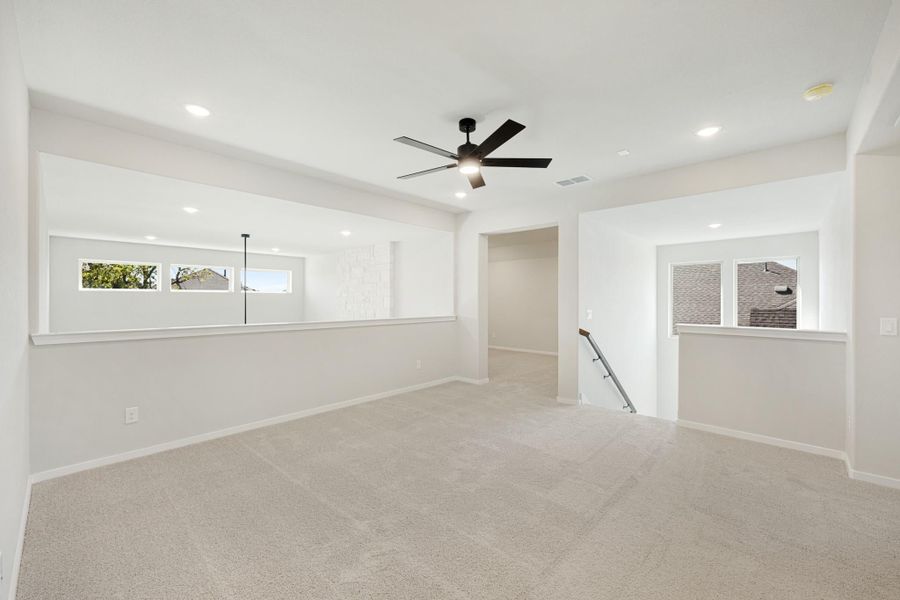 Open upstairs loft with carpet flooring, black ceiling fan, recessed lighting, and staircase opening with low half-walls and windows. Open upstairs loft with carpet flooring, black ceiling fan, recessed lighting, and staircase opening with low half-walls and windows.