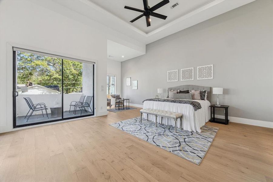 This first-floor primary bedroom features light wood flooring, high ceilings with recessed lighting and a modern fan, and sliding glass doors that open to a private outdoor balcony. The neutral walls and large windows enhance the bright and spacious ambiance.