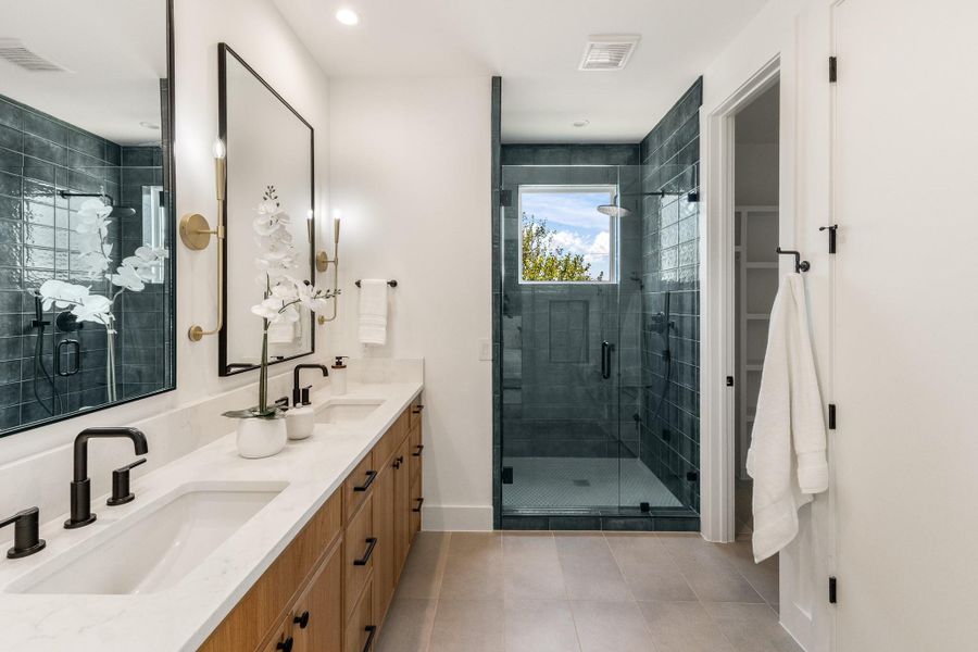 Dual vanities, warm wood cabinetry, quartz countertops, designer lighting, and a dramatic tiled walk-in shower create a serene, sophisticated retreat.