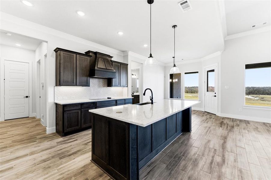 Kitchen featuring hanging light fixtures, light wood-type flooring, light stone counters, a kitchen island with sink, and ornamental molding