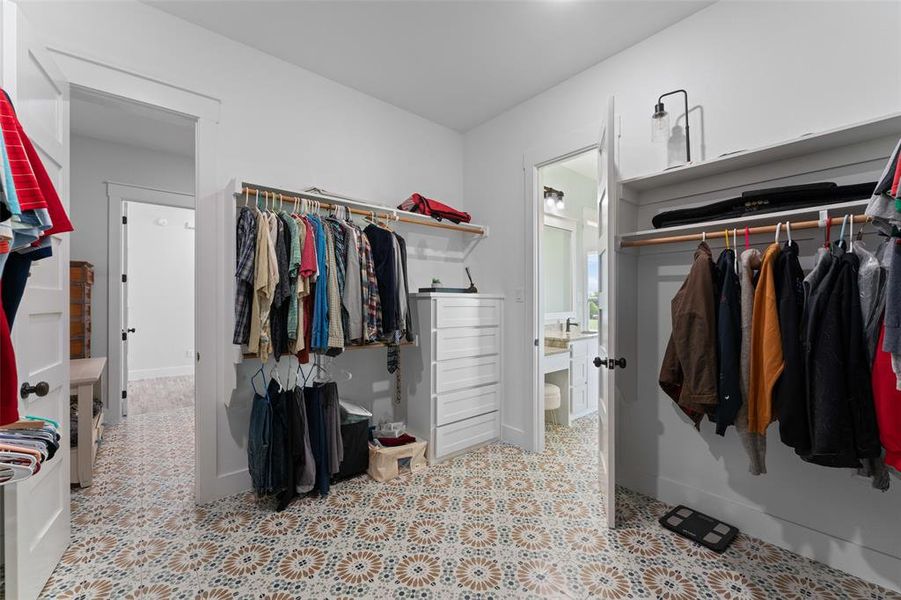 Spacious walk-in closet featuring patterned floor tiles, extensive built-in shelving, hanging rods, a white dresser, and a doorway opening to a bathroom with a vanity