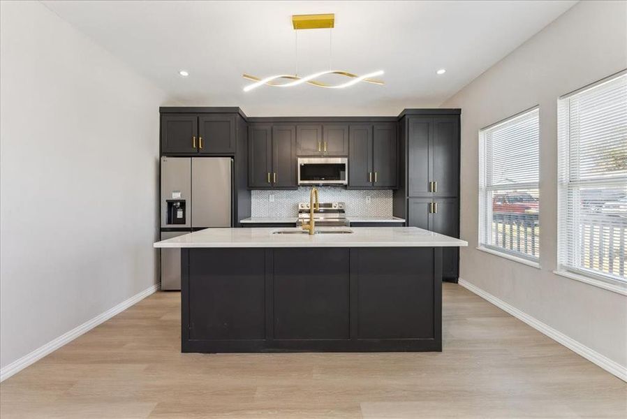 Kitchen with a kitchen island with sink, stainless steel appliances, a sink, baseboards, and decorative backsplash