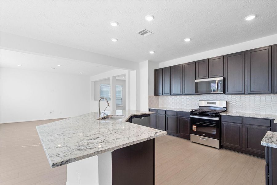This spacious kitchen boasts soaring ceilings, Woodmont Belmont cabinets in an Espresso stain, Granite Delicatus White countertops, stainless steel Whirlpool appliances, a modern Daltile backsplash, recessed lighting, an extended breakfast bar countertop, and a pantry—all overlooking the expansive family room and breakfast nook.