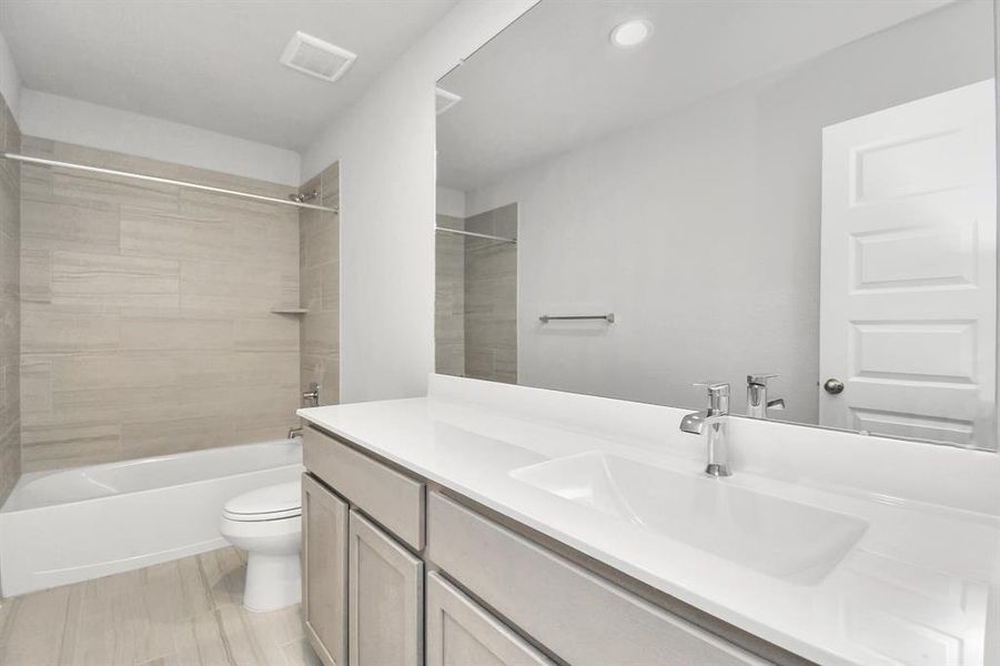 Secondary bathroom equipped with Bath/shower combo surrounded by tasteful tile. Light wood cabinets, coupled with a gorgeous countertop, elevate the space, cultivating a truly welcoming atmosphere.