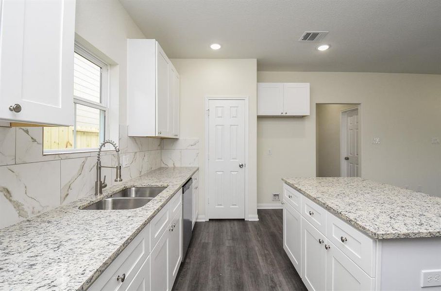 Modern kitchen interior with white cabinets, granite countertops, and stainless steel sink under a window, with neutral color walls and wood flooring.